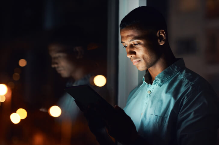 Man Stood By A Window At Night Looking At An Ipad